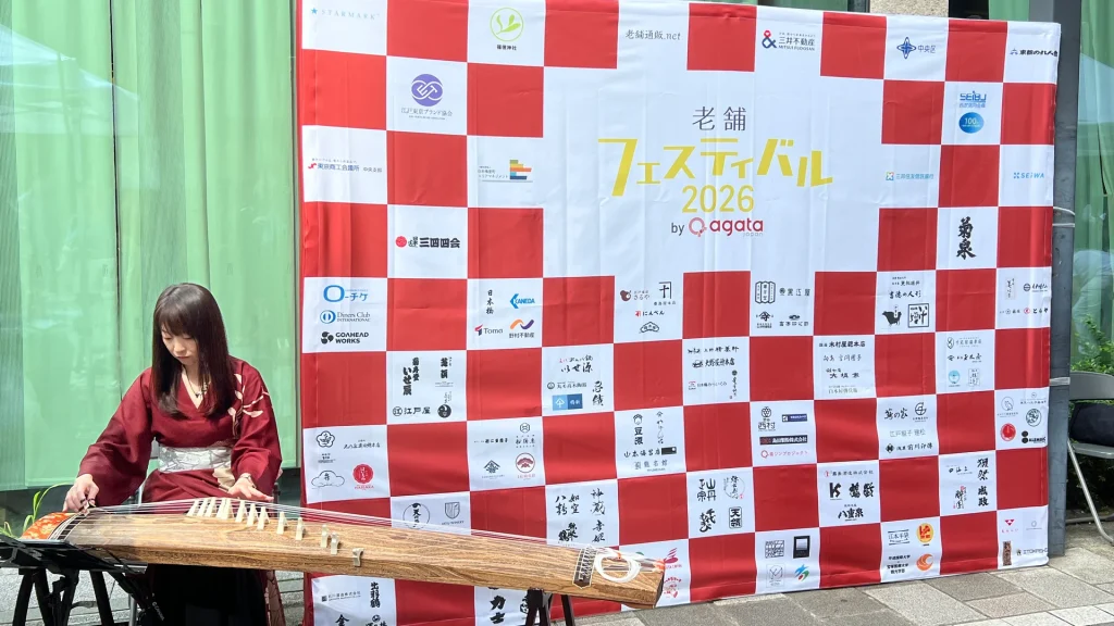 老舗フェスティバル2026のロゴボードの前で箏を演奏する様子
